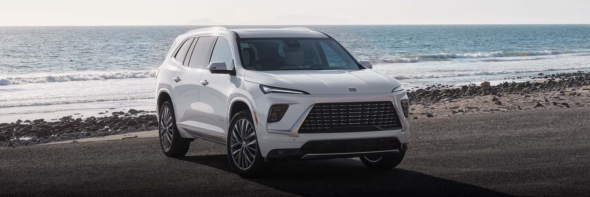 White SUV with black wheels parked near a rocky beach with blue ocean and small waves.